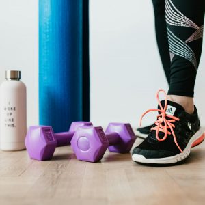 Crop sportswoman wearing sneakers and black leggings standing with sport mat on wooden floor near dumbbells and water bottle before exercising indoors