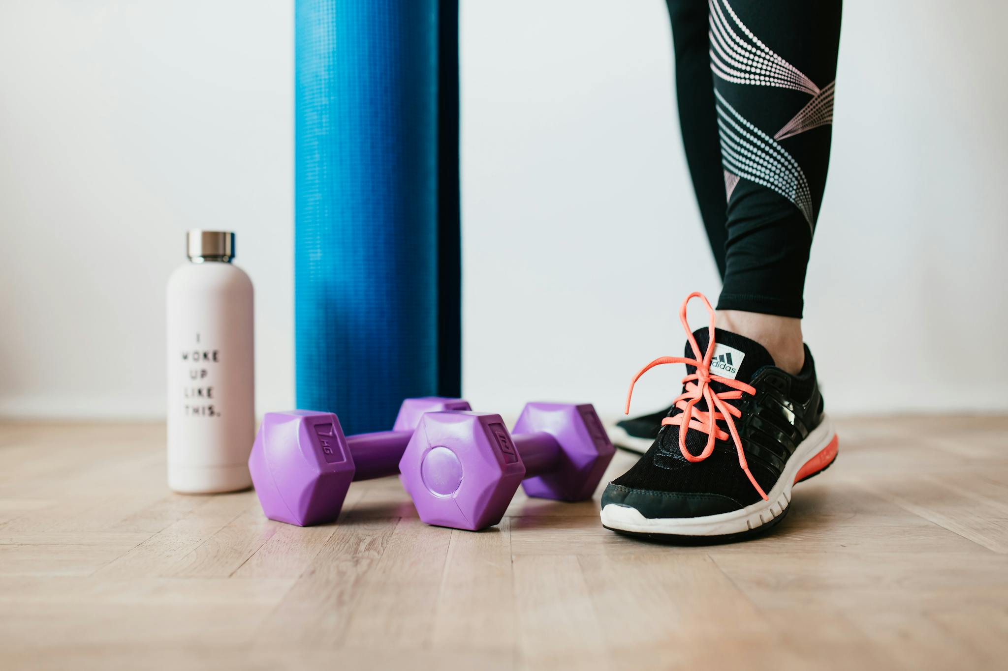 Crop sportswoman wearing sneakers and black leggings standing with sport mat on wooden floor near dumbbells and water bottle before exercising indoors