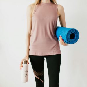 Faceless slim female athlete in sportswear standing with blue fitness mat and water bottle while preparing for indoors workout
