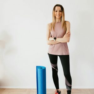 Full body female athlete wearing sportswear and sneakers standing against white wall with crossed arms near blue sport mat before yoga practice and looking at camera with smile