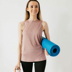 Positive young female athlete in sportswear standing isolated against white wall with blue yoga mat and water bottle while looking away with smile